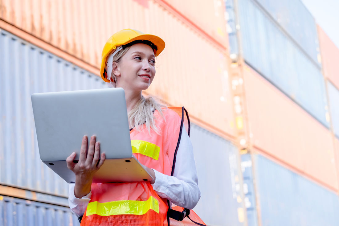 Woman cargo container technician or worker at a container terminal wearing an orange vest with neon yellow lines holding a laptop and looking to her left side