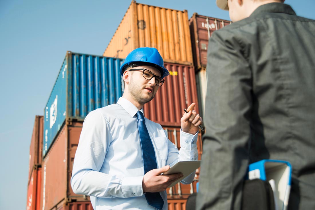 Male foreman wearing a blue hard hat, dress shirt, and blue tie speaking with a colleague at a container terminal while holding a notepad and a pen.