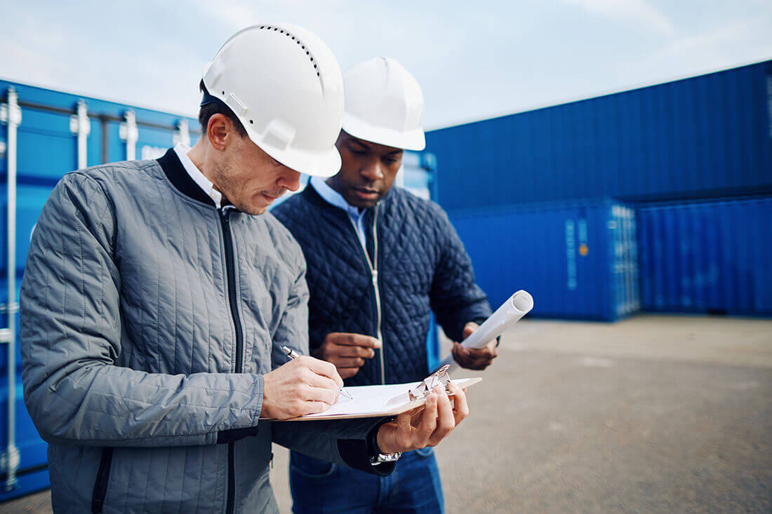 Two men wearing jackets and white hard hats discussing and writing in a terminal container yard.