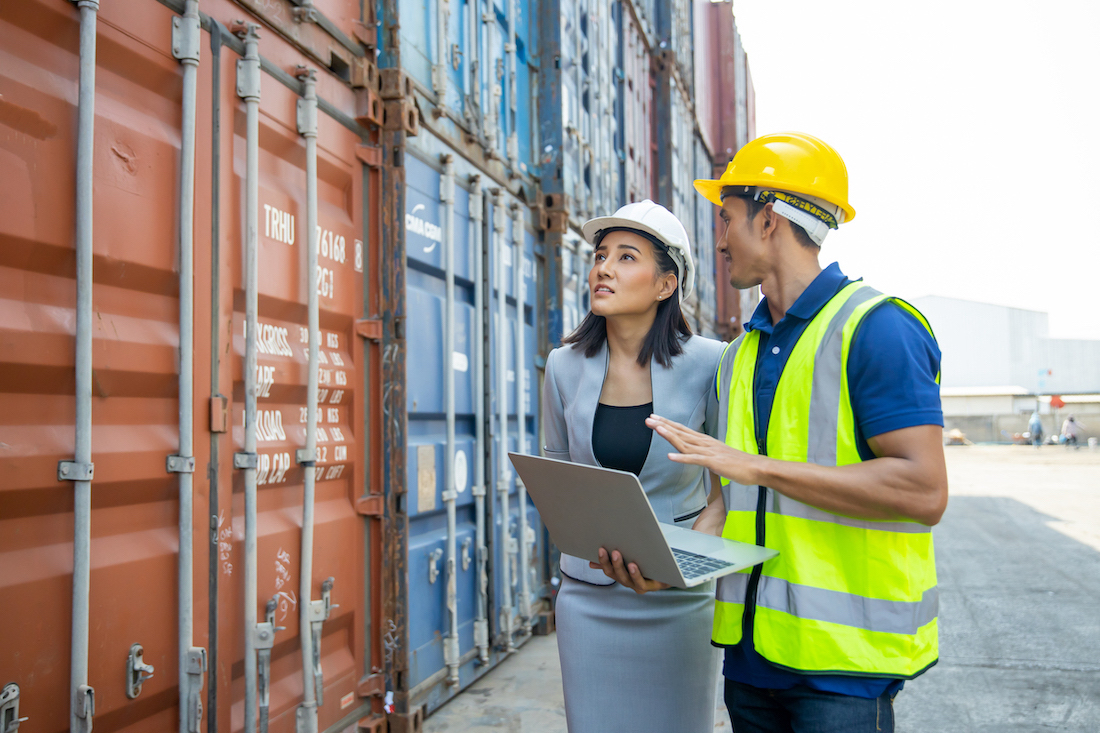 Business woman wearing a suit and a white hard hat talking to a port employee wearing a yellow hard hat and neon vest next to containers in a shipping yard