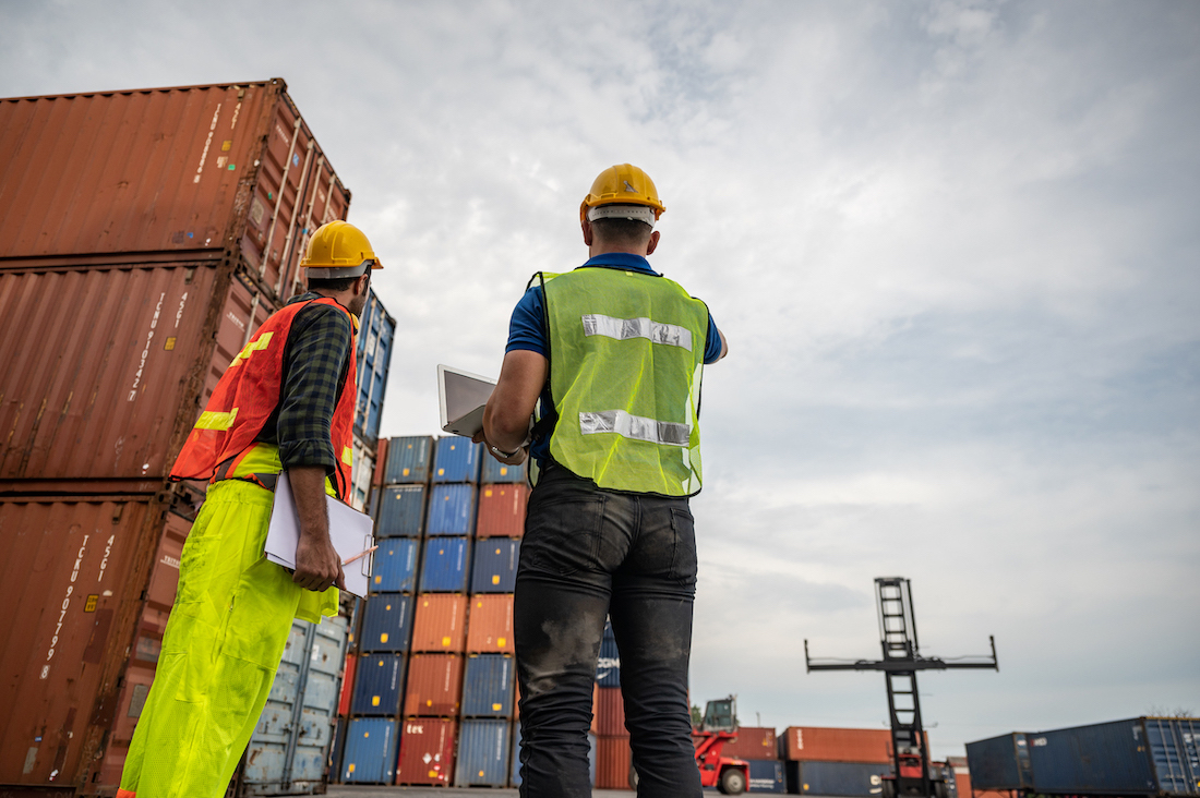 Two port employees wearing yellow hard hats and neon vests looking at a shipping container yard