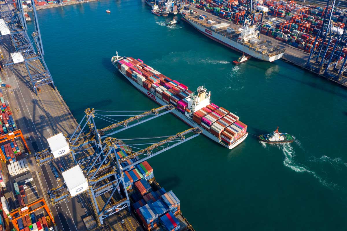 cargo ship docked at a busy shipping port, surrounded by colorful stacked containers and cranes, with a tugboat nearby in the water