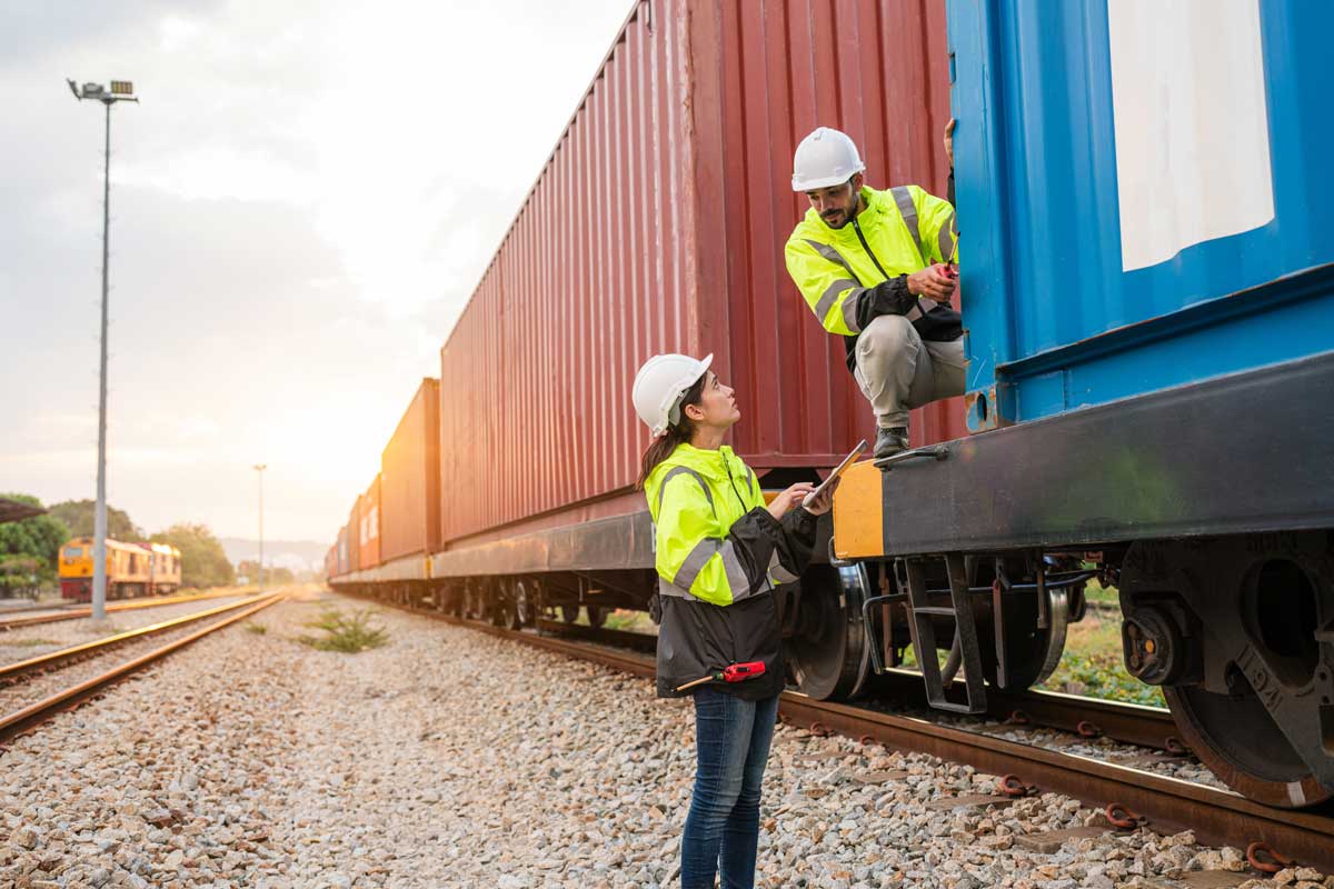 Two workers in high visibility safety gear involved in intermodal shipping operations, with one male worker seated on a freight train, communicating with a female worker standing below who is taking notes